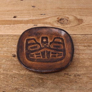 May include: A small, round, brown ceramic dish featuring a dark, embossed graphic of a stylised animal figure centred on its surface. The dish has a mottled, textured finish with darker brown tones highlighting the recessed design elements.