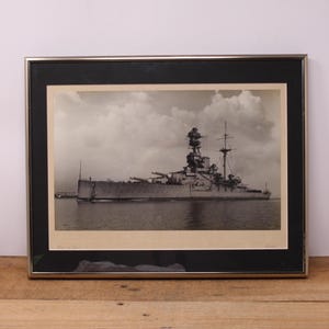 May include: A framed black and white photograph of a large battleship at sea. The ship is in the foreground and the sky is in the background. The ship has a large gun turret on the front and several smaller turrets on the sides. The ship is sailing on a calm sea with a few clouds in the sky.