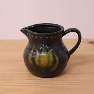 Peut inclure: Un pichet en céramique gris anthracite avec un corps arrondi, une anse incurvée et un bec verseur. L'extérieur est orné d'un motif floral peint à la main en forme de tulipe jaune et verte et d'une série de petites indentations sur le bord.