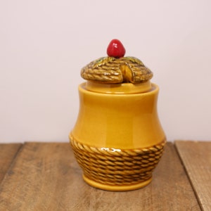 May include: A ceramic jar with a lid, featuring a basket weave design around the base and a nut-shaped lid with a red berry on top. The jar is a warm yellow color, and the lid is brown.