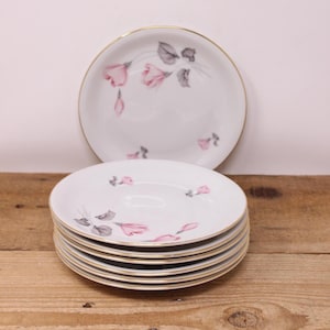 May include: A stack of white porcelain plates with a gold rim and a floral design in pink and gray. One plate is placed behind the stack. The plates are on a wooden surface.