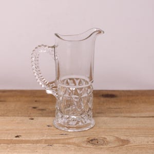 May include: A clear glass pitcher with a decorative handle and a diamond-patterned base. The pitcher has a curved spout and sits on a wooden surface. The handle has a twisted rope design.