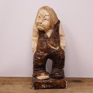 May include: A wooden figurine of a man with a grumpy expression, wearing a brown vest and pants. The figurine is standing on a wooden base with the word "remarkable" written on it.