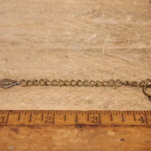 May include: A gold chain necklace with a lobster clasp closure. The chain is made of small, linked circles. The chain is measured against a wooden ruler.