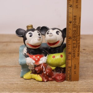 May include: Vintage ceramic figurine of Mickey Mouse and Minnie Mouse sitting on a blue bench. Mickey is wearing a red and white polka dot shirt and Minnie is wearing a yellow shirt with red polka dots. Both are wearing red shoes.