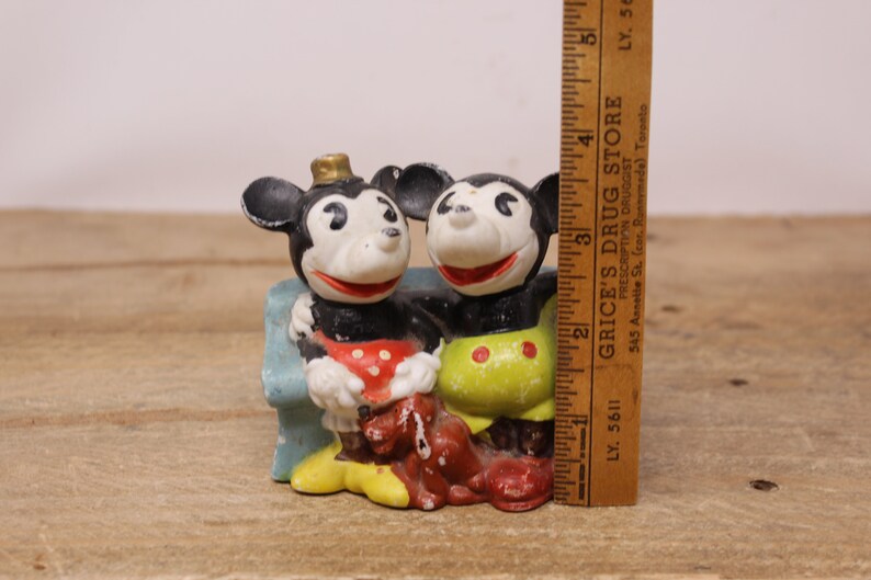 May include: Vintage ceramic figurine of Mickey Mouse and Minnie Mouse sitting on a blue bench. Mickey is wearing a red and white polka dot shirt and Minnie is wearing a yellow shirt with red polka dots. Both are wearing red shoes.