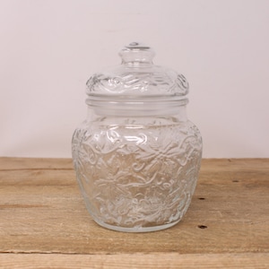 May include: A clear glass jar with a lid, both featuring an embossed floral design. The jar is on a wooden surface.