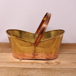 May include: A vintage brass and copper basket with a handle. The basket has a shiny gold finish with a copper band around the base and a copper handle. The basket is sitting on a wooden surface.