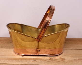 May include: A vintage brass and copper basket with a handle. The basket has a shiny gold finish with a copper band around the base and a copper handle. The basket is sitting on a wooden surface.