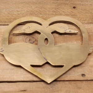 May include: A brass heart-shaped trivet featuring two stylised geese intertwined. The geese are facing opposite directions and their necks are intertwined.