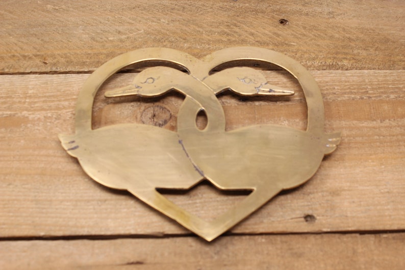 May include: A brass heart-shaped trivet featuring two stylised geese intertwined. The geese are facing opposite directions and their necks are intertwined.