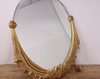 Può includere: Uno specchio da toeletta ovale dorato con una cornice ornata e arrotolata. Lo specchio è autoportante e ha una base decorativa.