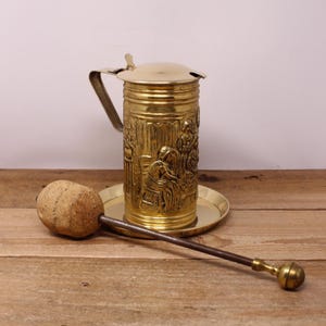 May include: A vintage brass beer stein with a lid, handle, and decorative relief carvings. The stein sits on a brass plate, with a cork and metal stopper resting nearby. The background is a wooden surface.