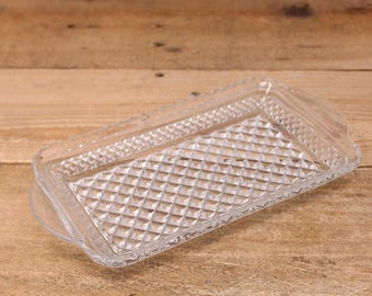 May include: A clear, rectangular glass tray with a diamond pattern on the base and a decorative border. The tray has two small handles and is set on a wooden surface. This vintage-style tray is suitable for serving or display.