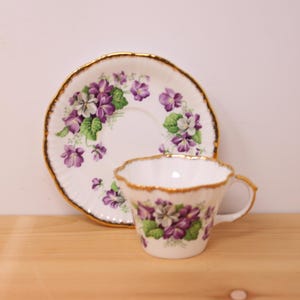 Peut inclure: Un ensemble tasse et soucoupe en porcelaine blanche orné d'un motif délicat de fleurs de violettes violettes et blanches avec des feuilles vertes. La tasse et la soucoupe ont des bords festonnés soulignés d'une fine finition dorée.