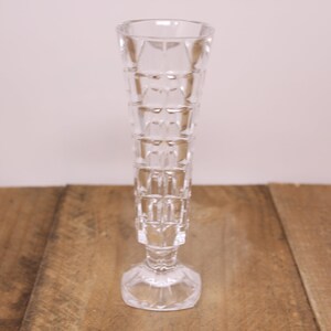 May include: A clear glass vase with a geometric pattern. The vase has a tall, slender body and a wide base.