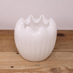May include: A white glass bowl with a ruffled edge. The bowl has a rounded shape with vertical ridges and a wavy top. The bowl is on a wooden surface.