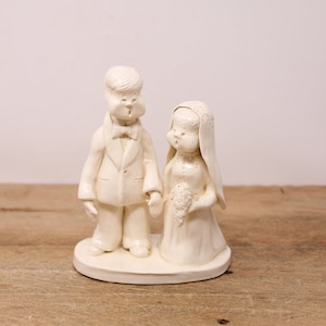 May include: White ceramic figurine of a bride and groom. The bride is wearing a veil and holding a bouquet of flowers. The groom is wearing a tuxedo and has a bow tie. The figurine is on a white base.