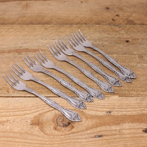 May include: Set of eight silver forks with ornate floral designs on the handles. The forks are arranged in a row on a wooden surface.