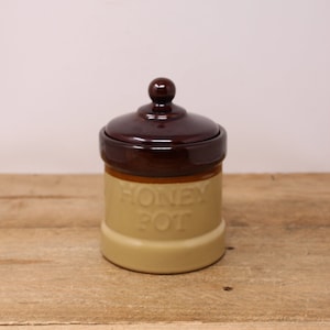 May include: A brown and beige ceramic honey pot with a lid. The pot has the words "Honey Pot" embossed on the front.