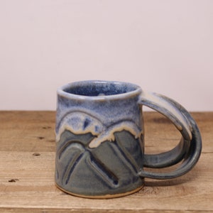 May include: A blue and white ceramic mug with a mountain design and a double handle.