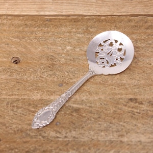 May include: A silver serving spoon with an ornate, pierced design on the circular head. The handle features detailed floral patterns. The spoon is made of a shiny, reflective metal and rests on a wooden surface.