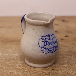 May include: A small, white ceramic jug with a blue handle and a blue inscription that reads "A'1813 Jack Amsterdam".