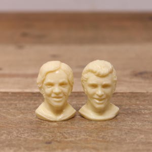 May include: Two off-white ceramic figurines of men's heads. The figurines are detailed with facial features and hair.