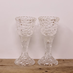 May include: Two clear glass candle holders with a textured, bubble-like design. The holders have a tall, slender stem and a wide, round base.