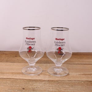 May include: Two clear Pilsner beer glasses with a silver rim. Each glass has the words "Moninger Ratsherrn Pilsener" printed in red and a small red devil graphic. The glasses are on a wooden surface.