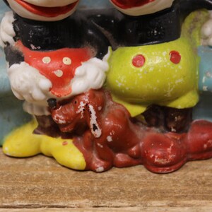 May include: Vintage ceramic figurine of Mickey Mouse and Minnie Mouse sitting on a blue bench with a brown dog. Mickey is wearing red shorts and Minnie is wearing yellow shorts. The figurine is in good condition with some paint loss.