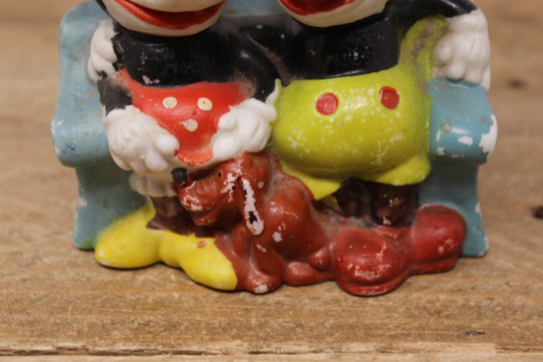 May include: Vintage ceramic figurine of Mickey Mouse and Minnie Mouse sitting on a blue bench with a brown dog. Mickey is wearing red shorts and Minnie is wearing yellow shorts. The figurine is in good condition with some paint loss.