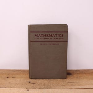May include: An old book, titled "Mathematics for Technical Schools", with an olive green cover. The title and author's name are printed in maroon. The book is standing on a wooden surface.