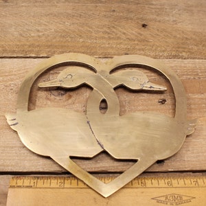 May include: A brass heart-shaped hot pad featuring two stylised geese facing each other. The geese are intertwined in a heart shape.