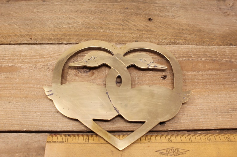 May include: A brass heart-shaped hot pad featuring two stylised geese facing each other. The geese are intertwined in a heart shape.