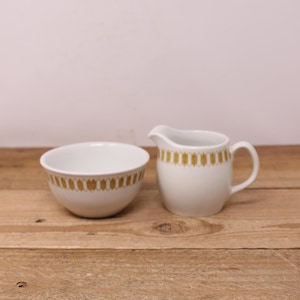 May include: A white ceramic bowl and creamer set with a gold geometric pattern. The bowl is round, and the creamer has a handle and spout. The set is displayed on a wooden surface. This vintage set is perfect for serving cream and sugar.
