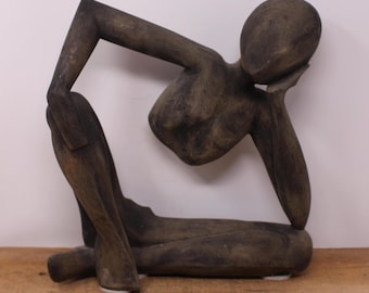 May include: A dark brown wooden sculpture of a person in a seated pose, with one arm bent and resting on the head. The figure is seated with legs bent and the other arm extended. The sculpture is on a wooden surface.