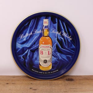 May include: A vintage round serving tray with a blue and gold rim. The tray features a bottle of "Bulloch Lade Scotch Whisky" against a blue velvet backdrop. The bottle has a red and white label with the letters "B L".