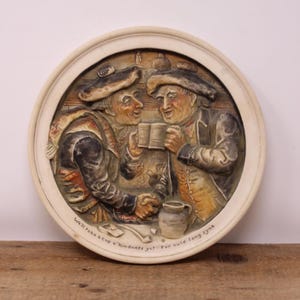May include: A white ceramic wall plaque with a raised relief image of two men in period clothing, drinking from mugs. The plaque has a brown wooden frame and the text "We'll take a cup o' kindness yet" For build lang tyne.