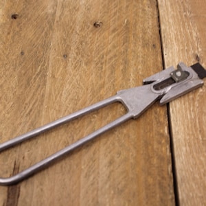 May include: A silver metal tool with a handle and a blade. The blade has a serrated edge and a small hole in the center. The tool is lying on a wooden surface.