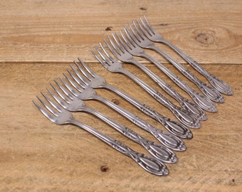 May include: A set of eight silver forks with ornate handles. The forks are arranged in a row, with the handles pointing to the right. The forks are on a wooden surface.