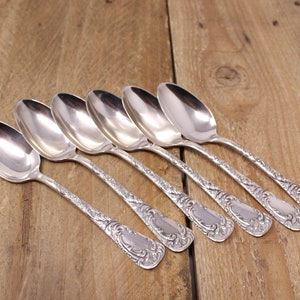 May include: Six silver spoons with ornate handles, arranged in a row on a wooden surface.