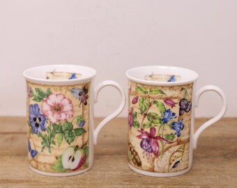 May include: Two white ceramic mugs with floral designs. One mug features pink and blue flowers, an apple slice, and butterflies. The other mug displays purple and blue flowers, shells, and berries. Both mugs have a vintage, botanical theme.