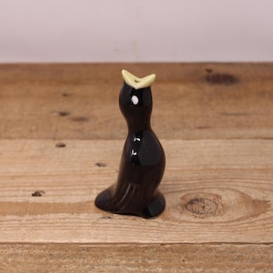 May include: A small, black ceramic figurine of a bird with a yellow beak and a white dot on its head. The bird is standing upright on a wooden surface. The figurine has a glossy finish and a simple, elegant design.