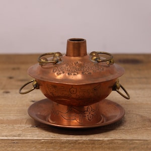 May include: A small copper hot pot with a decorative floral design and three brass handles. The pot sits on a matching copper plate.