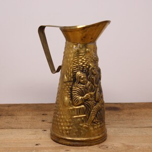 May include: A vintage brass pitcher with a handle and a detailed embossed design. The pitcher features a scene with figures and a textured surface. The handle is a simple, curved design. The pitcher is likely used for serving beverages.