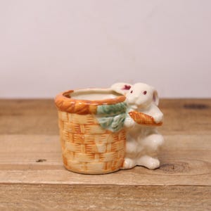 Peut inclure: Une figurine de lapin en céramique blanche tenant une carotte et regardant hors d'un pot de fleurs en osier brun.