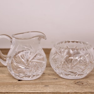 May include: A clear glass creamer jug and sugar bowl set. The jug has a handle and a spout, while the bowl is round with a scalloped rim. Both pieces feature a starburst pattern. The set is displayed on a wooden surface.