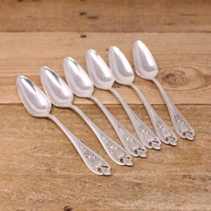 May include: Six silver spoons arranged diagonally on a wooden surface. The spoons have oval bowls and ornate detailing on the handles. The spoons are reflective, and the wooden surface has a warm, natural tone.