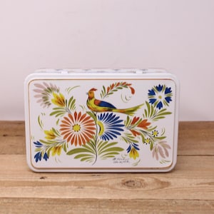 May include: White metal box with a colorful floral and bird design. The artwork features a yellow, orange, blue, and green bird perched among orange, blue, and yellow flowers. The box has a gold trim and a hinged lid.
